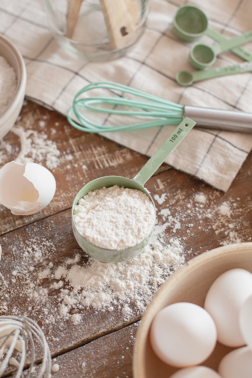 Green measuring cup filled with flour sits on a flour-covered wooden surface. Cracked egg shells, a whisk, and tea towel are placed around it.