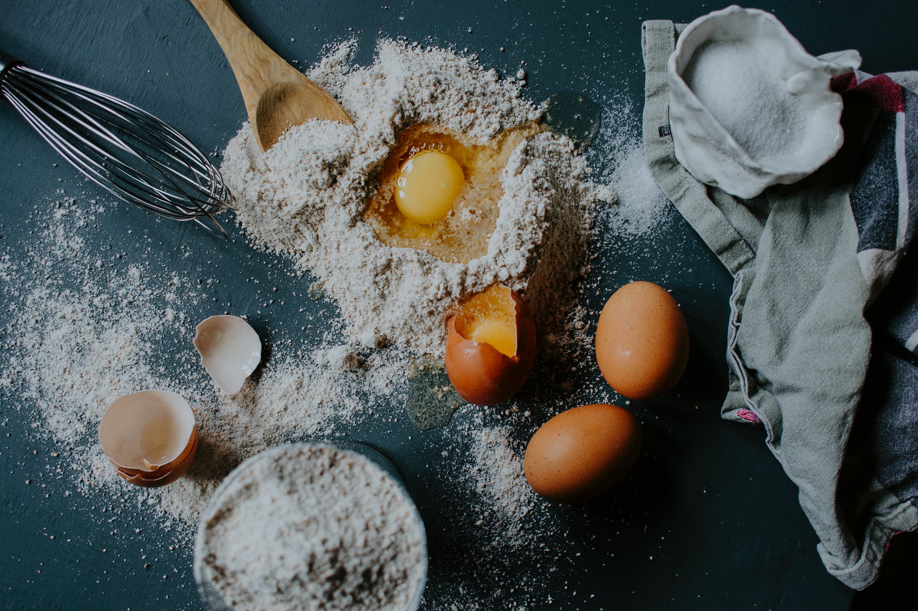 A flour-covered surface with eggs, cracked egg shells, a tea towel, and bowl of flour. 
